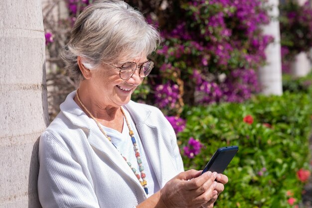 Ritratto di bella donna anziana che si appoggia su una pianta mentre si utilizza lo smartphone in una soleggiata giornata estiva