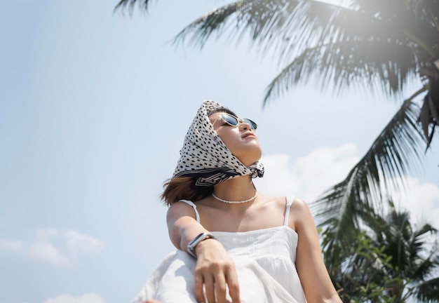 Ritratto di bella donna asiatica felice in camicia bianca casual che indossa occhiali da sole e sciarpa per capelli guardando il sole in spiaggia sotto le palme da cocco e lo sfondo del cielo blu in estate