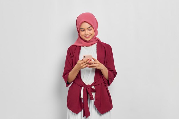 Ritratto di bella donna asiatica sorridente in camicia casual e hijab utilizzando un telefono cellulare, digitando messaggi sms isolati su sfondo bianco. Concetto di stile di vita religioso della gente