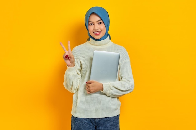 Ritratto di bella donna asiatica sorridente in maglione bianco e hijab che tiene laptop e mostra il segno di pace con le dita isolate su sfondo giallo persone concetto di stile di vita religioso