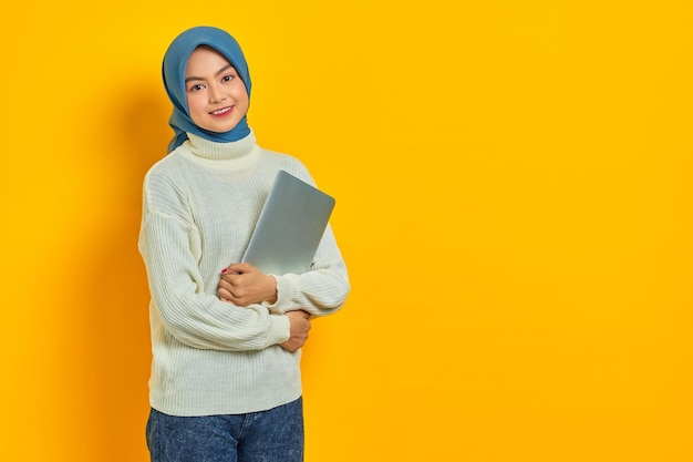 Ritratto di bella donna asiatica sorridente in maglione bianco e hijab in possesso di un laptop e guardando la fotocamera isolata su sfondo giallo persone concetto di stile di vita religioso