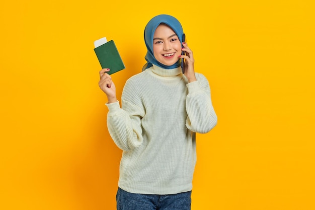 Ritratto di bella donna asiatica sorridente in maglione bianco parlando sul cellulare e tenendo il passaporto isolato su sfondo giallo Summer Trip