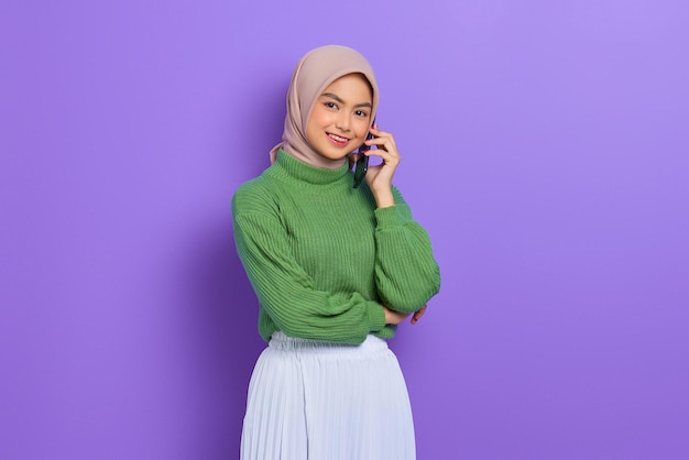 Ritratto di bella donna asiatica sorridente in maglione verde e hijab che parla al telefono cellulare isolato su sfondo viola