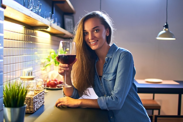 Ritratto di bella donna bevente felice sorridente casuale giovane carina che tiene un bicchiere di vino rosso e seduto al tavolo nella cucina in stile loft a casa