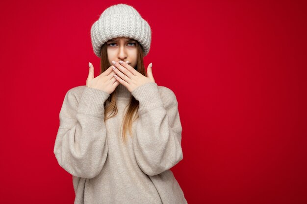 Ritratto di bella donna bionda scura adulta insoddisfatta arrabbiata con emozioni sincere che indossa un maglione caldo e cappello lavorato a maglia isolato su sfondo rosso con spazio libero e che copre la bocca con le mani.