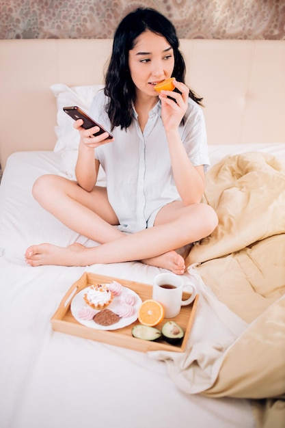 Ritratto di bella donna bruna che indossa indumenti da notte seduta con le gambe incrociate a letto guardando il cellulare in mano, facendo una gustosa colazione, mangiando arancia, avacado nel suo accogliente appartamento.