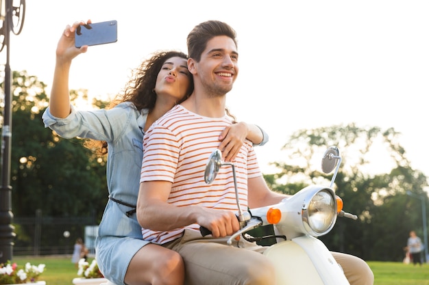 Ritratto di bella donna che cattura selfie sullo smartphone, mentre guida una moto attraverso una strada cittadina insieme al suo fidanzato