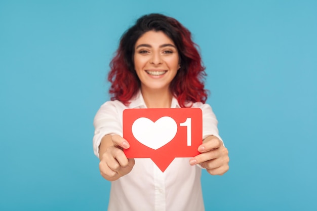 Ritratto di bella donna con capelli rossi fantasia che mostra il cuore dei social media come icona, simbolo di popolarità in internet, che consiglia di premere il pulsante. tiro in studio indoor isolato su sfondo blu