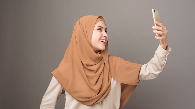 Ritratto di bella donna con hijab tramite cellulare su sfondo grigio