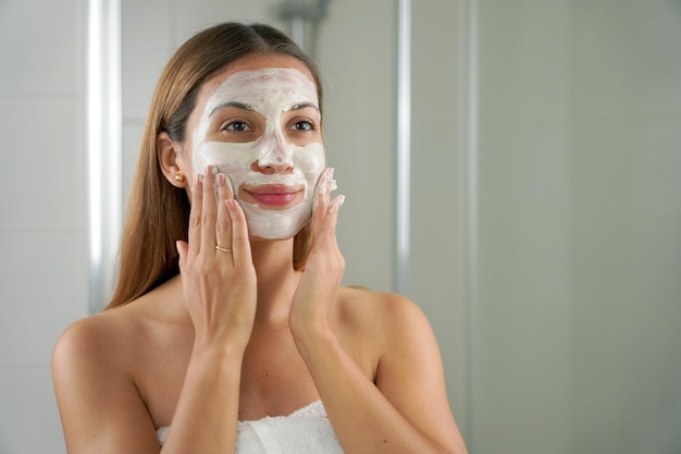 Ritratto di bella donna con una maschera di argilla sul viso che si guarda allo specchio in bagno