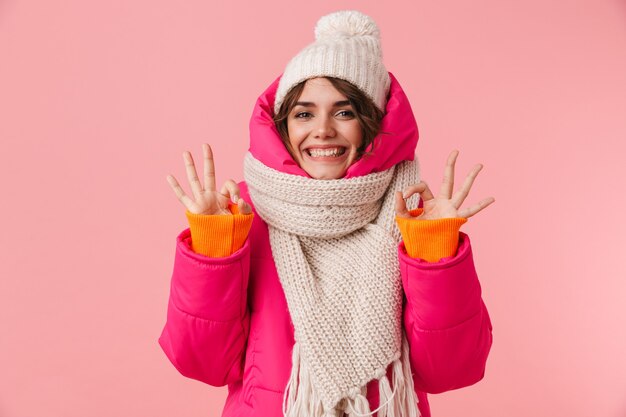 Ritratto di bella donna contentissima in vestiti caldi che gesticolano segno ok e sorridente isolato sul rosa