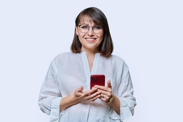 Ritratto di bella donna di mezza età felice con il telefono che guarda l'obbiettivo su sfondo bianco studio Donna matura sorridente in occhiali con acconciatura trucco Tecnologia stile di vita business anni '40 persone