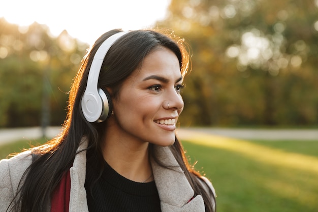 Ritratto di bella donna felice che indossa cappotto ascoltando musica con le cuffie e sorridendo mentre si cammina nel parco autunnale