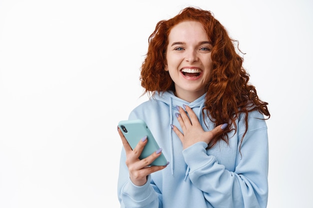 Ritratto di bella donna felice con i capelli rossi ricci, ridendo dopo aver letto il messaggio sullo smartphone, tenendo il telefono cellulare e sorridendo su bianco