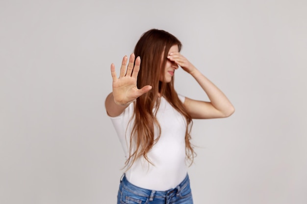 Ritratto di bella donna impaurita che chiude gli occhi con il palmo e mostra il gesto della mano di arresto girando il viso non vuole vedere indossare una maglietta bianca Indoor studio girato isolato su sfondo grigio