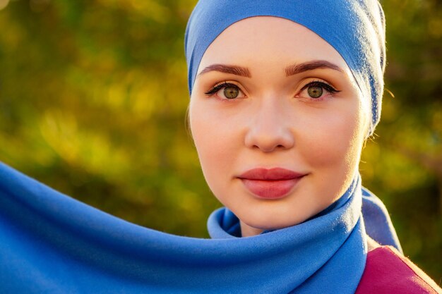 Ritratto di bella donna musulmana con gli occhi verdi che indossa una sciarpa blu con il viso chiuso coperto con alberi forestali di sfondo avelo nel parco