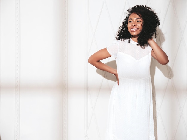 Ritratto di bella donna nera con l'acconciatura afro riccioli Modello sorridente vestito in abito estivo bianco Femmina spensierata sexy in posa vicino al muro in studio Abbronzata e allegra in una giornata di sole