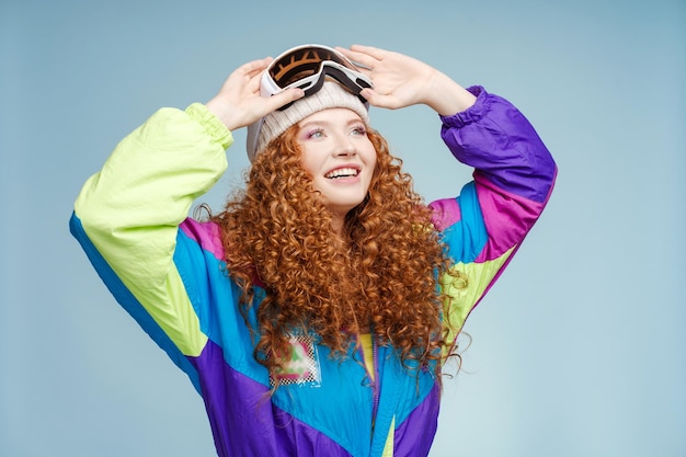 Ritratto di bella donna sorridente dai capelli ricci che indossa cappello e occhiali protettivi da sci tuta da sci vintage distogliendo lo sguardo isolato su sfondo blu Concetto di pubblicità di viaggio invernale