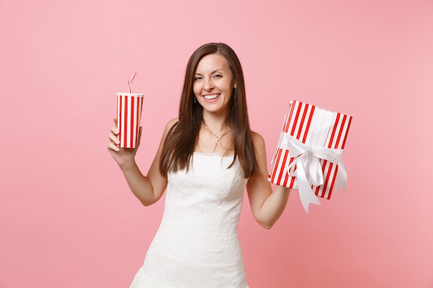 Ritratto di bella donna sorridente in vestito bianco che tiene scatola rossa con regalo, presente tazza plactic con cola o soda