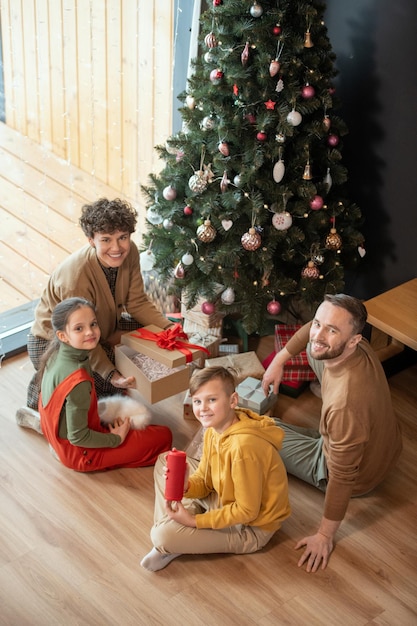 Ritratto di bella famiglia caucasica positiva seduta con regali sul pavimento contro l'albero di natale...