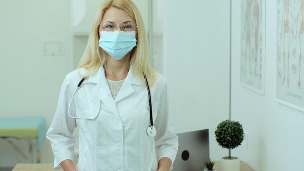 Ritratto di bella femmina caucasica medico professionista in maschera medica in piedi nel suo ufficio della clinica sanitaria. Concetto di medicina, persone e malattie infettive.