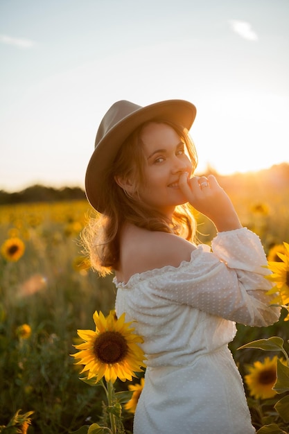 Ritratto di bella giovane donna 33 anni in cappello in campo di girasoli al tramonto Modello felice in abito bianco la sera d'estate in natura Caldo