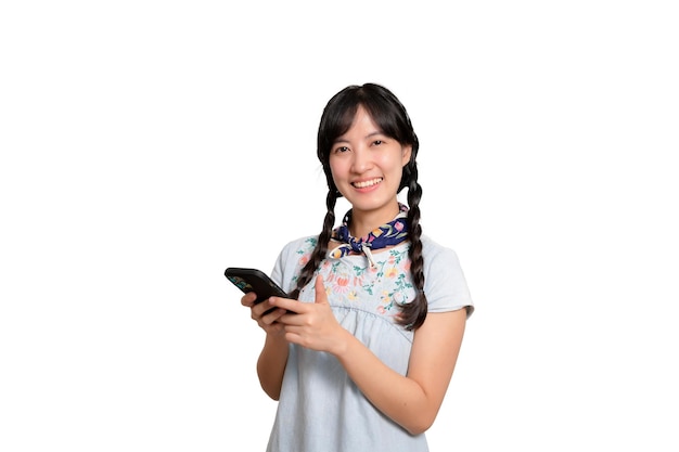 Ritratto di bella giovane donna asiatica felice in vestito dal denim facendo uso di uno smartphone sul colpo bianco dello studio del fondo