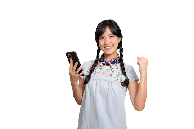 Ritratto di bella giovane donna asiatica felice in vestito dal denim facendo uso di uno smartphone sul colpo bianco dello studio del fondo