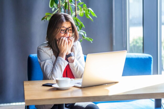 Ritratto di bella giovane donna bruna alla moda nervosa con gli occhiali seduto e guardando il monitor del laptop, mordendosi le unghie e preoccupandosi o facendosi prendere dal panico. girato in studio al coperto, caffè, sfondo dell'ufficio.