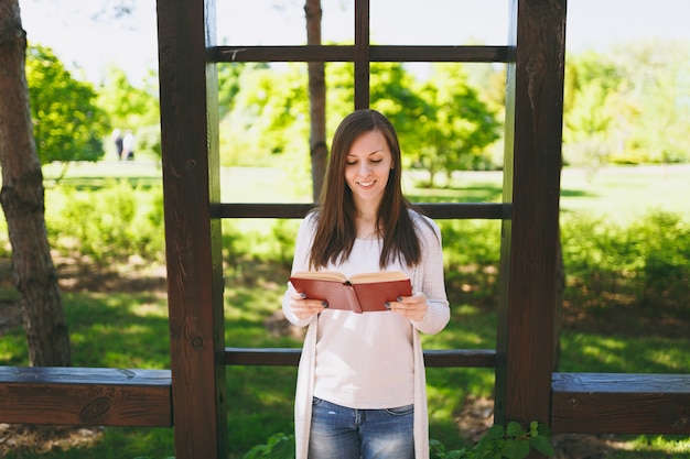 Ritratto di bella giovane donna calma e pacifica che indossa abiti casual leggeri, jeans, rilassante, libro di lettura. Donna sorridente che riposa nel parco cittadino in strada all'aperto sulla natura primaverile. Concetto di stile di vita.