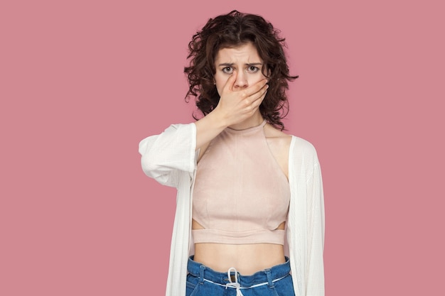 Ritratto di bella giovane donna castana preoccupata scioccata con l'acconciatura riccia in stile casual in piedi, coprendosi la bocca e guardando la fotocamera. studio al coperto colpo isolato su sfondo rosa.