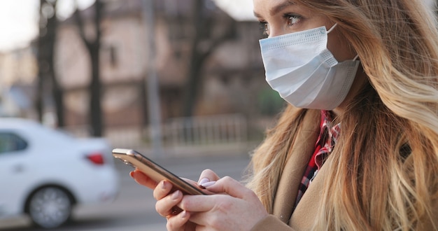 Ritratto di bella giovane donna caucasica nella mascherina medica usando il suo smartphone all'aperto. Messaggio mandante un sms della ragazza graziosa sul telefono durante la pandemia alla via. Concetto pandemico, epidemia di coronavirus.