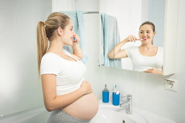 Ritratto di bella giovane donna che lava i denti allo specchio in bagno