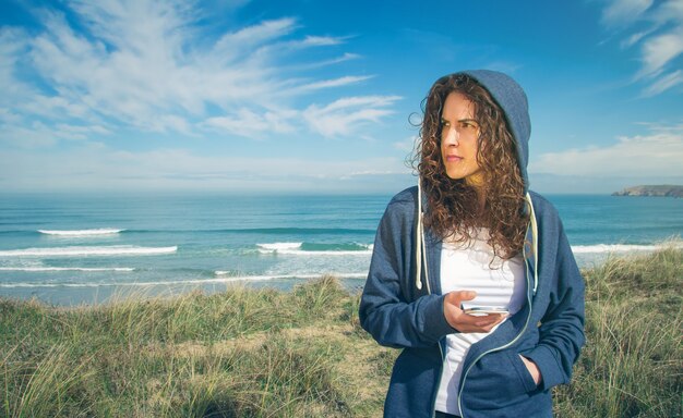 Ritratto di bella giovane donna con felpa con cappuccio blu e abbigliamento sportivo che tiene il suo smartphone su uno sfondo di cielo blu nuvoloso cloud