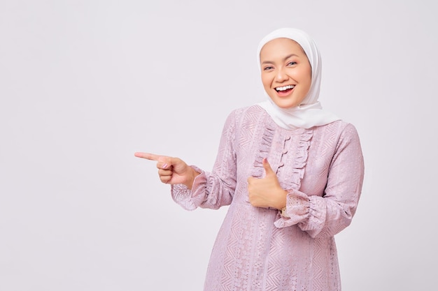 Ritratto di bella giovane donna musulmana asiatica sorridente che indossa l'hijab e il vestito viola che punta il dito da parte lo spazio vuoto e dà il pollice in alto segno gesto isolato su sfondo bianco studio