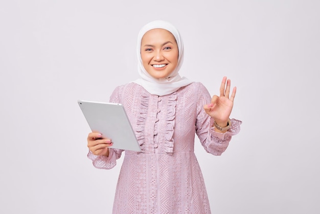 Ritratto di bella giovane donna musulmana asiatica sorridente che indossa l'hijab e il vestito viola che tiene la tavoletta digitale e mostra il segno ok ok isolato su sfondo bianco studio