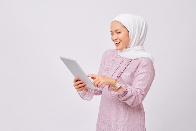 Ritratto di bella giovane donna musulmana asiatica sorridente che indossa l'hijab e vestito viola che tiene la tavoletta digitale isolata su sfondo bianco dello studio