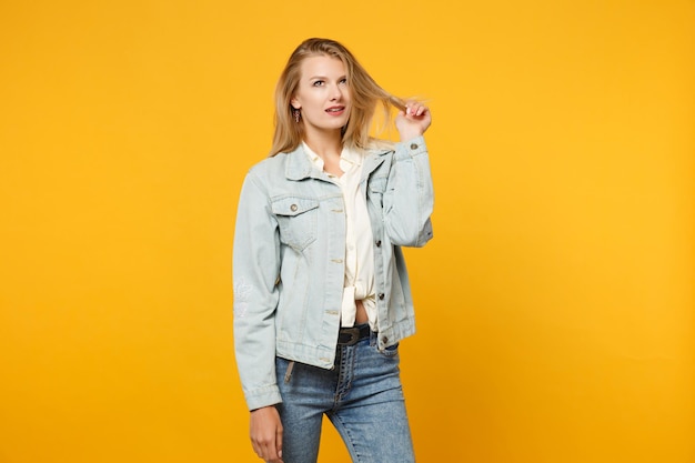 Ritratto di bella giovane donna pensosa in abiti casual in denim che guarda in alto e tiene i capelli isolati su uno sfondo di parete arancione giallo brillante in studio. Concetto di stile di vita delle persone. Mock up spazio di copia.