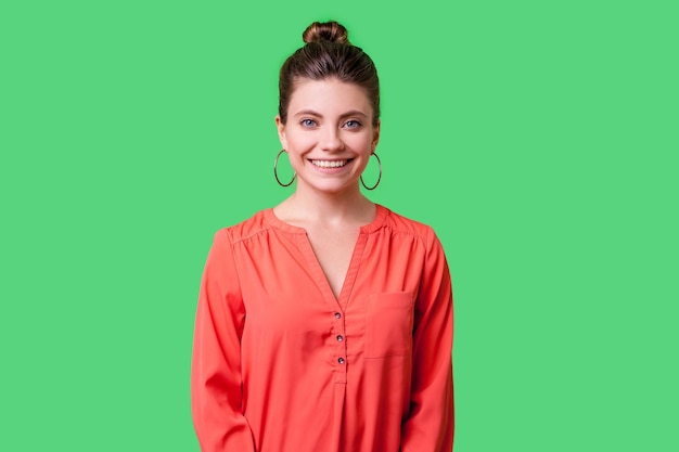 Ritratto di bella giovane donna positiva con acconciatura panino grandi orecchini e in camicetta rossa in piedi guardando la fotocamera con sorriso affascinante a trentadue denti in studio indoor girato isolato su sfondo verde