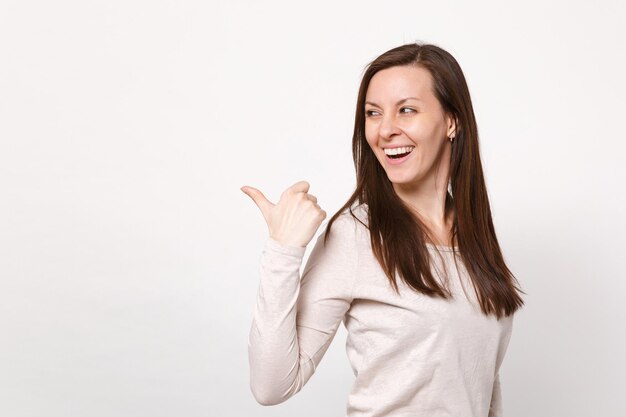 Ritratto di bella giovane donna ridente in abiti leggeri che guarda, puntando il pollice da parte isolato su sfondo bianco muro in studio. Persone sincere emozioni, concetto di stile di vita. Mock up spazio di copia.