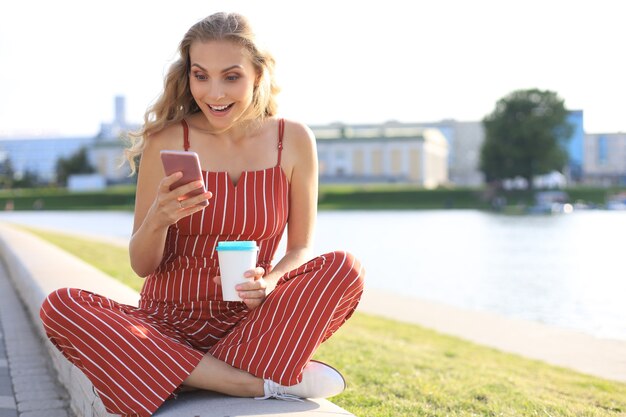 Ritratto di bella giovane donna seduta sulla riva del fiume con le gambe incrociate durante il giorno d'estate, sorridente e utilizzando lo smartphone.