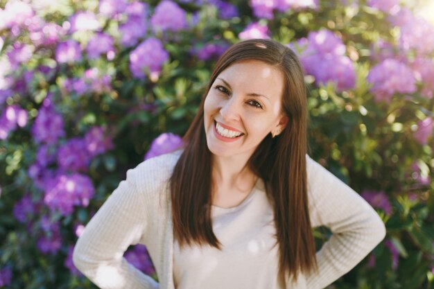 Ritratto di bella giovane donna sensuale dei capelli del brunette con il sorriso affascinante che indossa vestiti casuali leggeri. Donna sorridente vicino al cespuglio fiorito nel parco cittadino all'aperto sulla natura primaverile. Concetto di stile di vita.