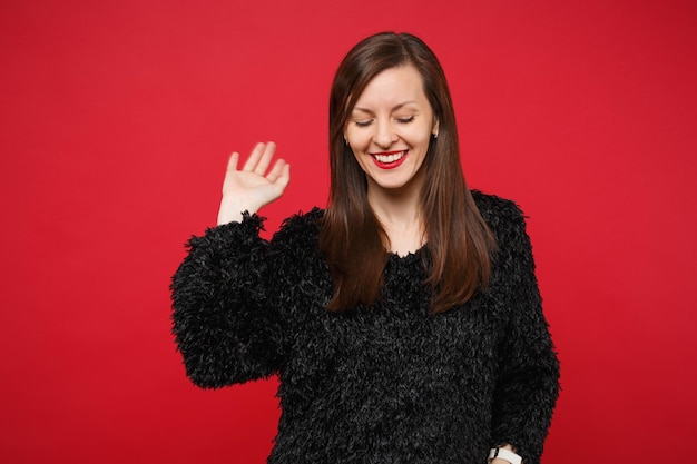 Ritratto di bella giovane donna sorridente in maglione di pelliccia nera che guarda giù agitando la mano isolata su fondo rosso brillante della parete in studio. Concetto di stile di vita di emozioni sincere della gente. Mock up copia spazio.