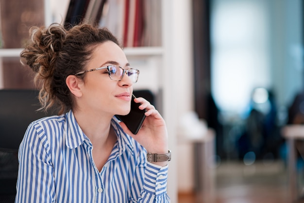 Ritratto di bella giovane imprenditrice con gli occhiali seduto al posto di lavoro e parlando sul telefono cellulare