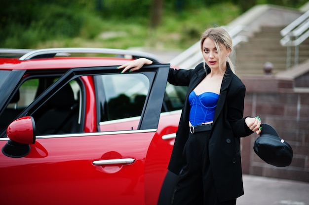 Ritratto di bella modella bionda sexy moda donna in berretto e in corsetto tutto nero, blu, con trucco luminoso vicino a city car rossa.