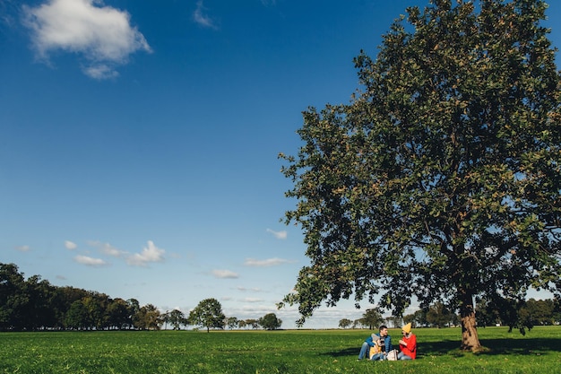 Ritratto di bella natura e famiglia che fa un picnic sullo sfondo si siede sotto il grande albero Cielo azzurro ed erba verde Incantevole clima autunnale e paesaggi Tre persone bevono il tè all'aperto sul prato