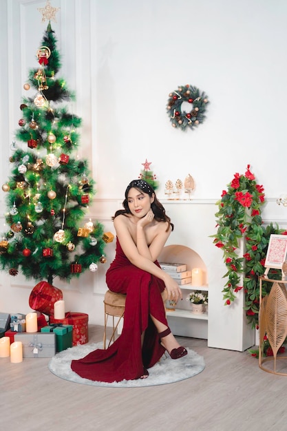 Ritratto di bella ragazza accogliente sedersi sorridente indossare abito rosso nel soggiorno di Natale decorato al chiuso