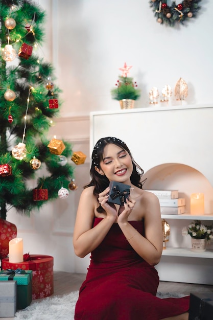 Ritratto di bella ragazza accogliente sedersi tenendo il regalo di Natale sorridente indossare abito rosso nel soggiorno di Natale decorato al chiuso