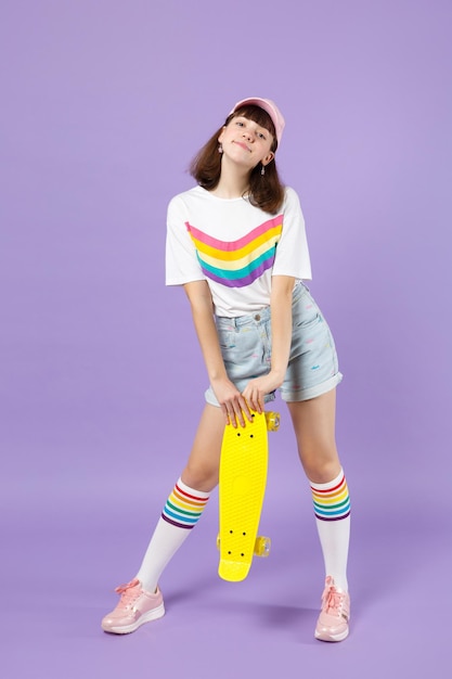 Ritratto di bella ragazza adolescente in abiti vivaci in piedi, con in mano uno skateboard giallo isolato su sfondo viola pastello in studio. Persone sincere emozioni concetto di stile di vita. Mock up spazio di copia.