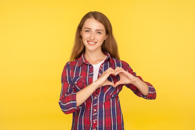 Ritratto di bella ragazza adorabile di zenzero con un sorriso gentile e in camicia a scacchi che mostra la forma del cuore con le mani, gesticolando amore, carità e concetto di speranza. girato in studio isolato su sfondo giallo